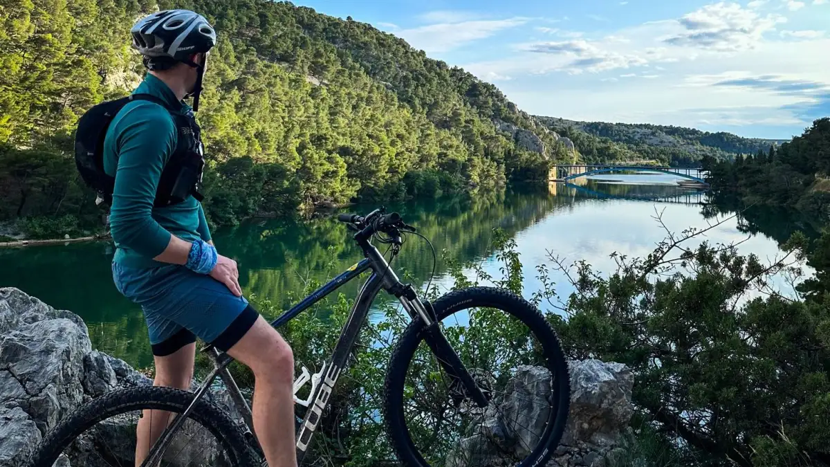 Cyclist in teal attire resting and admiring the view of a tranquil river and lush greenery in Krka National Park, with his mountain bike beside him on a rocky outcrop