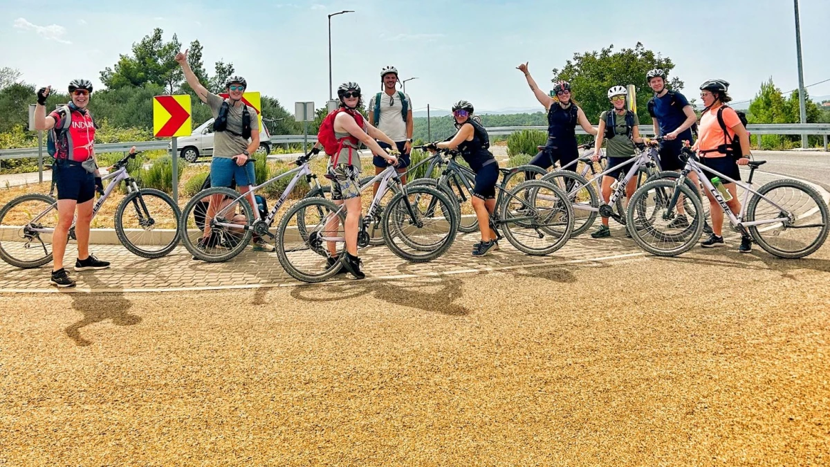 Nine happy cyclists pose with their bikes on a paved path, some with raised arms and thumbs up, wearing casual gear and helmets, with greenery in the background on a sunny day.