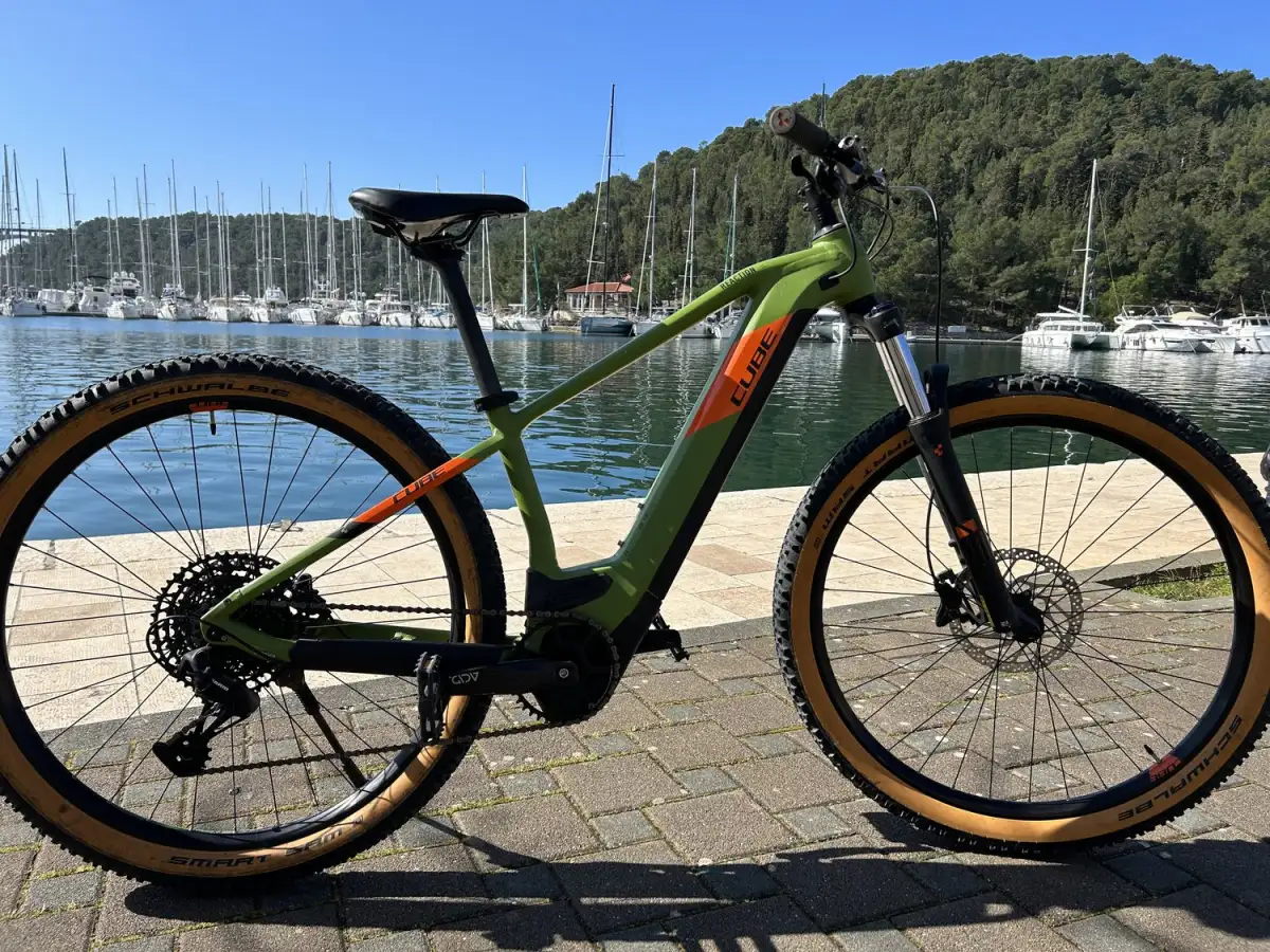 Modern green and orange e-bike positioned on a sunny marina promenade with a view of moored sailboats and a forested hill in the background