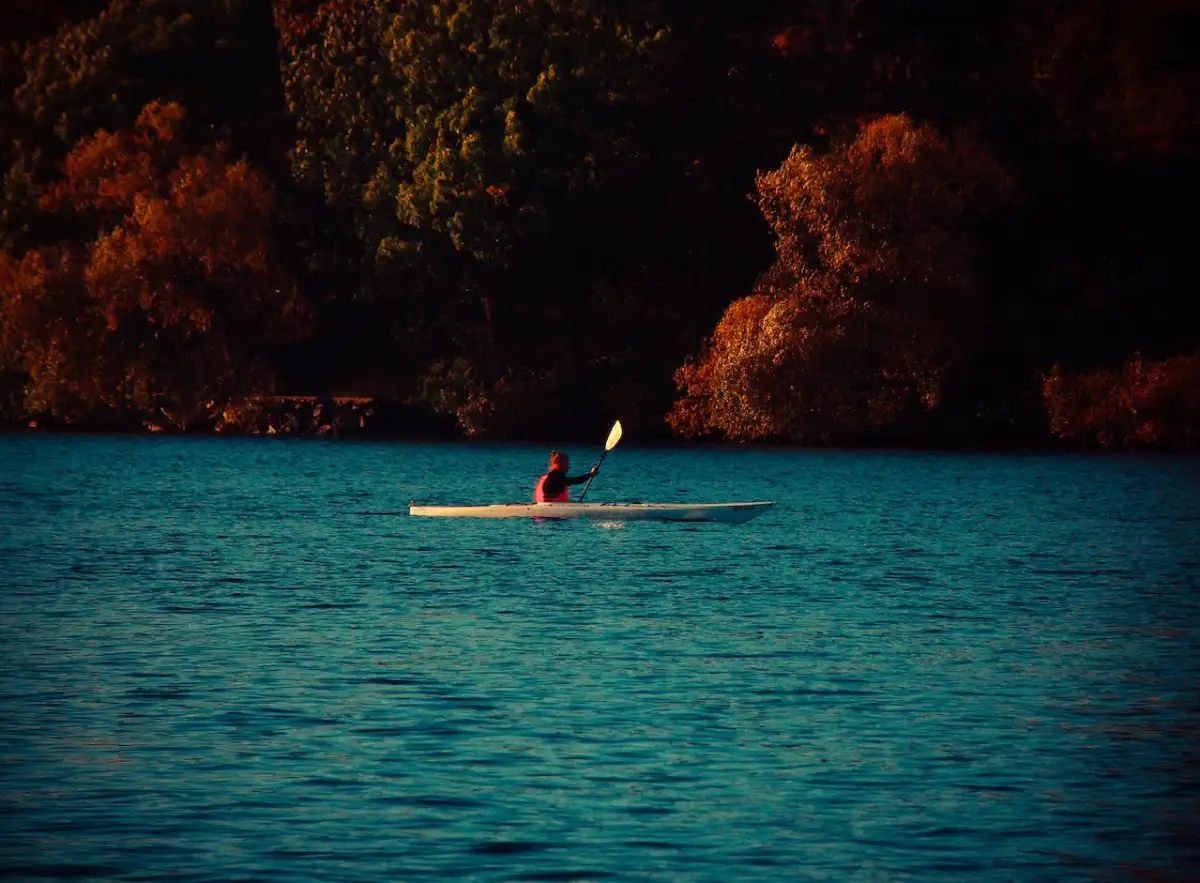 Solitary kayaker paddling on a tranquil blue lake with autumn-colored trees reflecting on the water surface during sunset