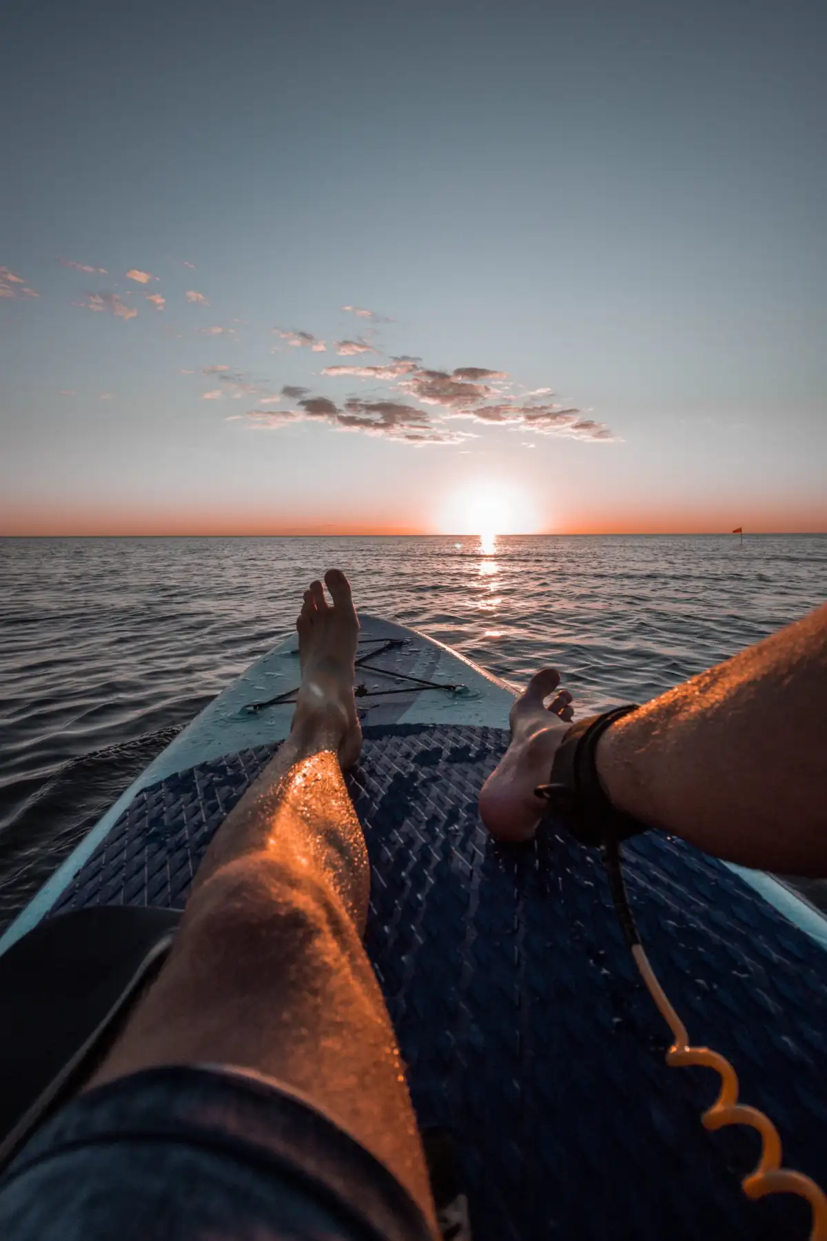 First-person view of a person's legs on a stand-up paddleboard, gliding over calm waters towards a beautiful sunset horizon.