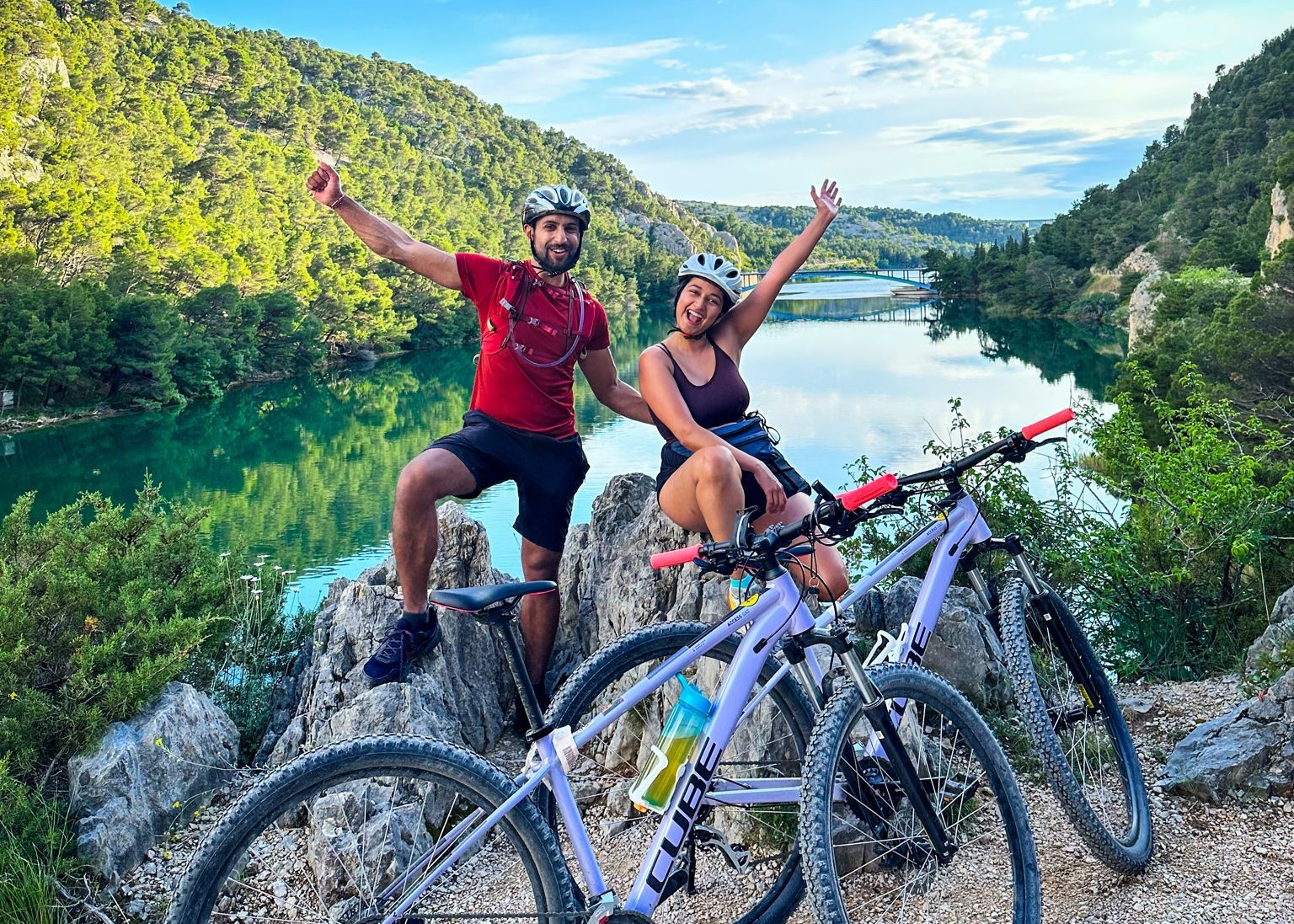 Two happy mountain bikers posing triumphantly atop a rocky outcrop with their bicycles, overlooking a serene lake surrounded by lush greenery in the Krka National park