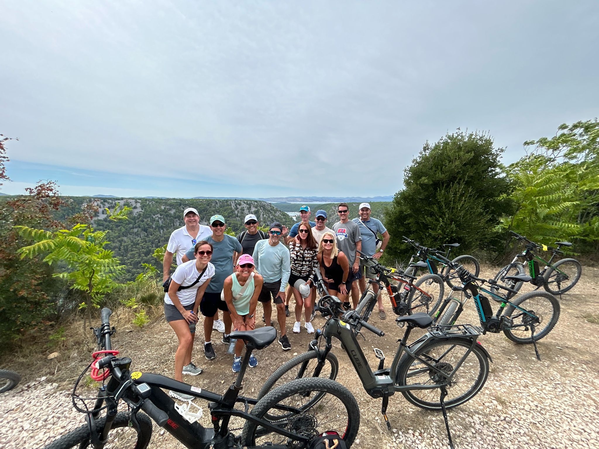 Group of happy bikers posing with their mountain bikes on a gravel trail overlooking a scenic landscape with lush hills and a distant water view