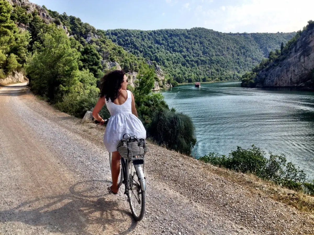 Woman in a white dress biking along a gravel path beside the serene blue waters of Krka National Park with lush green hills in the background and a boat visible in the distance.